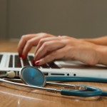Eine Frauenhand tippt auf einem Laptop. Daneben liegt ein Stethoskop.